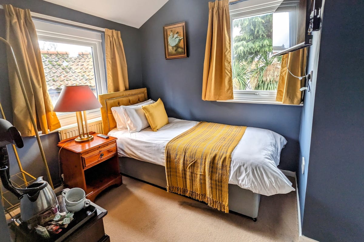 Quarto com cama, mesa de cabeceira, abajur, janela com cortinas e radiador.