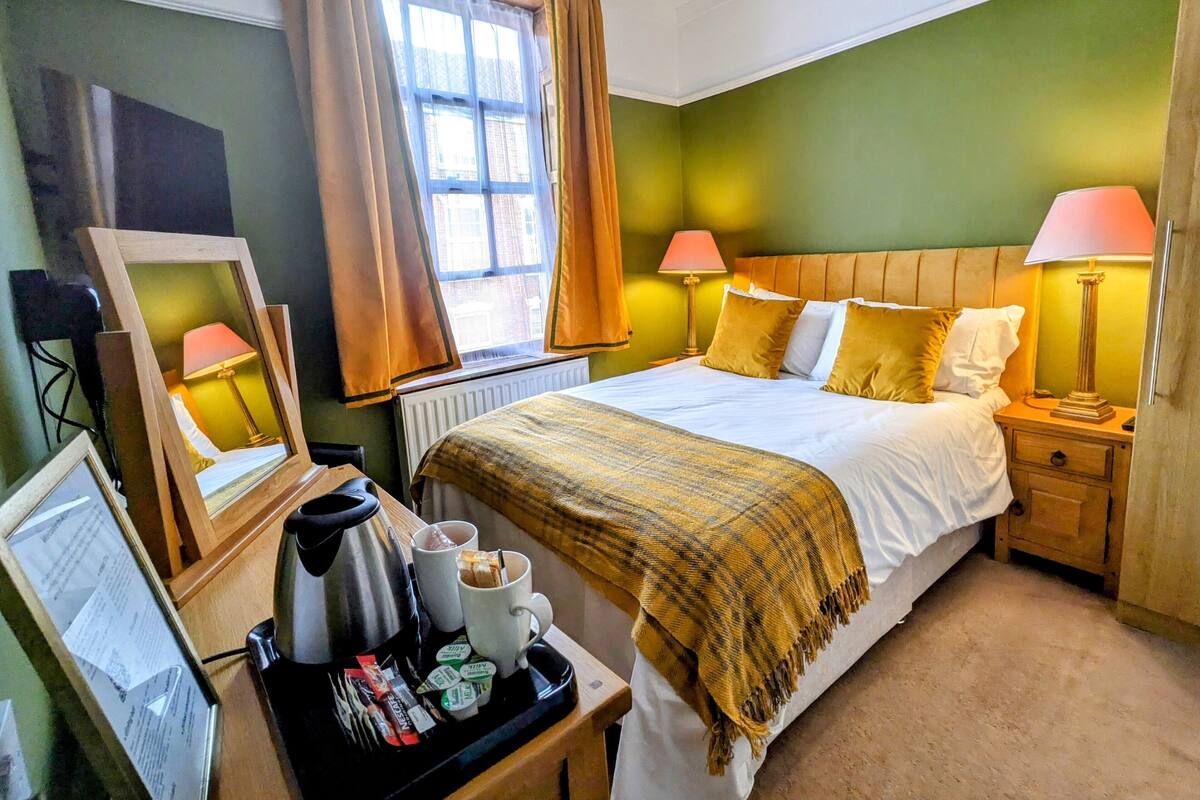 Quarto de hotel com cama, luminárias de cabeceira, escrivaninha com cafeteira e janela com cortinas.
