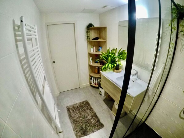 A bathroom with a white radiator, a wooden shelf with toiletries, a sink with a mirror, and a small rug on the floor. - Charming and Cosy rooms Nantes (Nantes)
