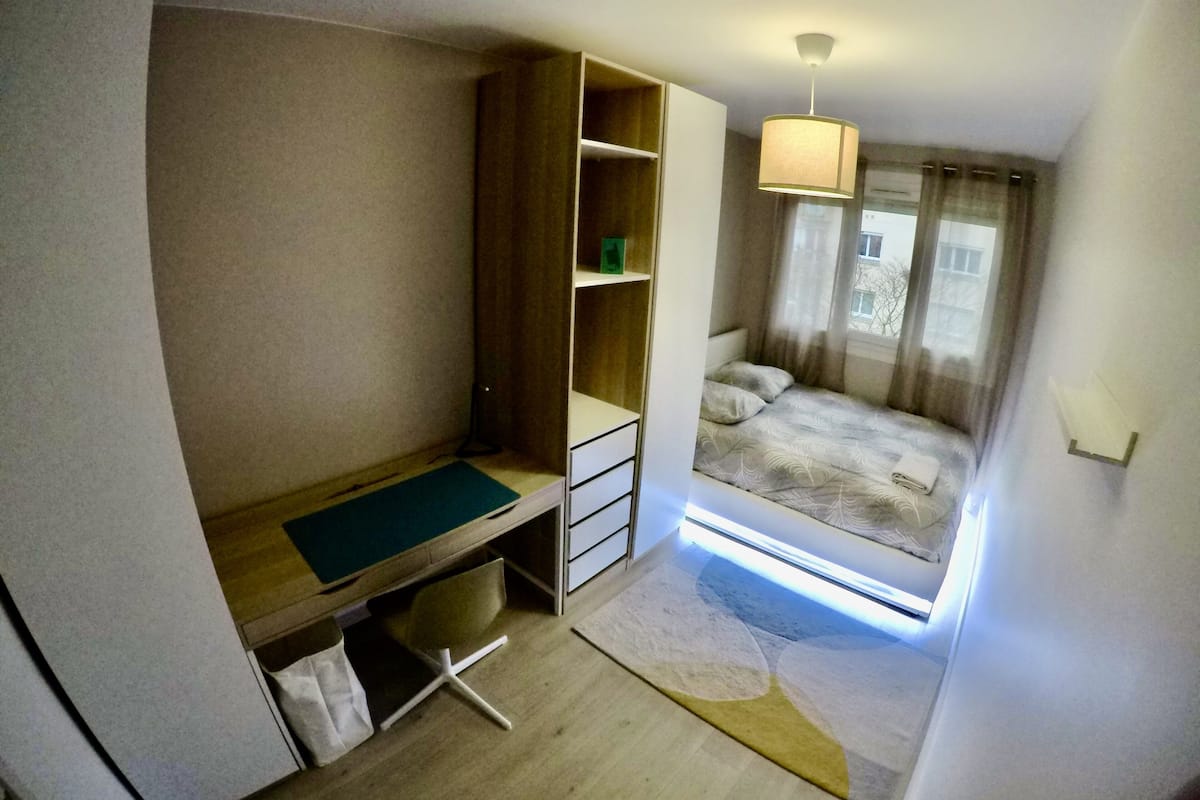 Una habitación compacta con una cama individual, un escritorio con silla, un armario y una ventana pequeña con cortinas.
