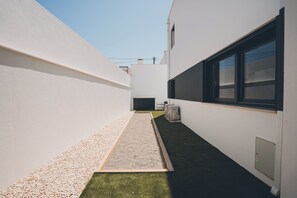 A narrow outdoor corridor with a gravel path and artificial grass, bordered by white walls and a building with large windows.
