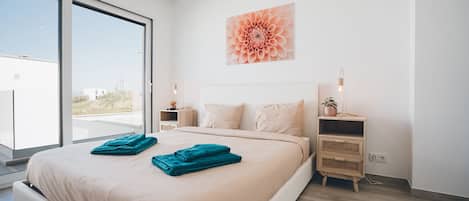 Une chambre moderne avec un grand lit, deux tables de chevet, une fenêtre offrant une belle vue et une œuvre d’art florale accrochée au mur.