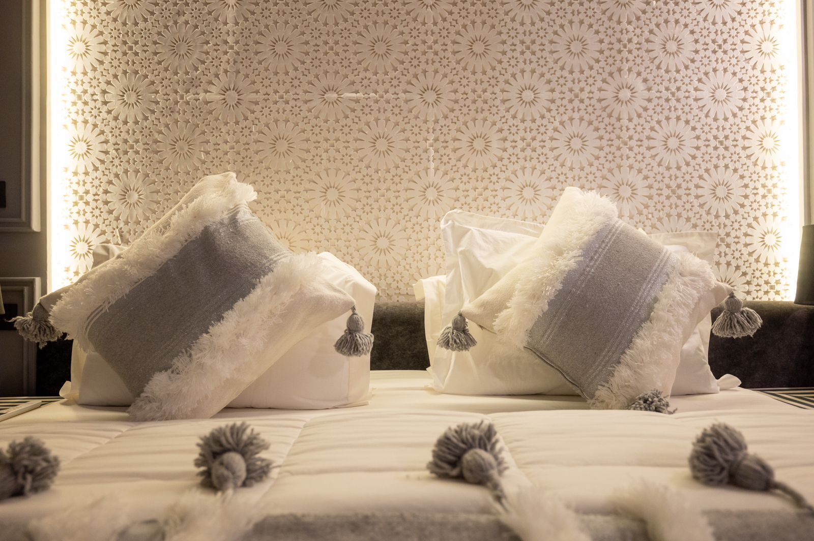 A hotel room with a bed featuring a white and gray striped blanket and a decorative pillow with tassels. The wall has a floral pattern.