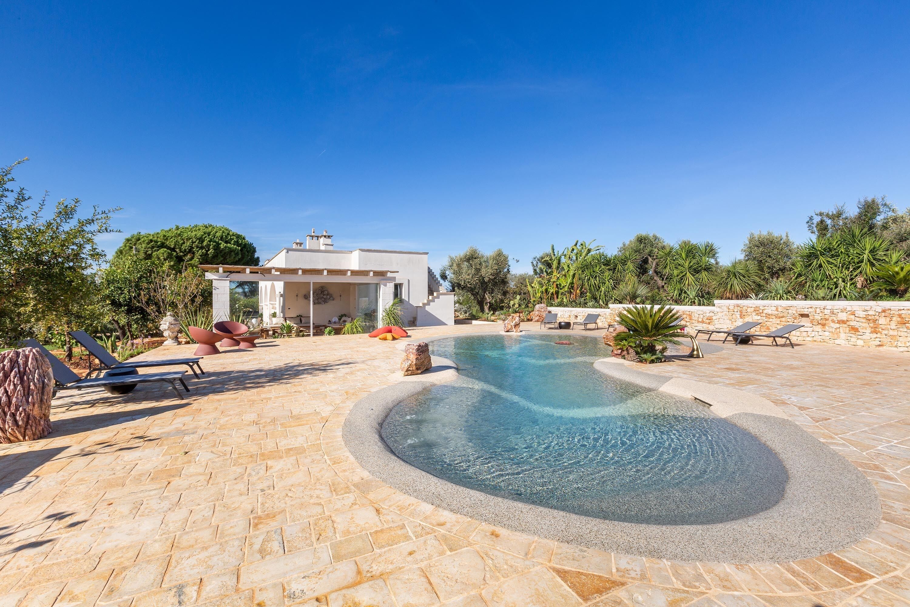 Une piscine à bordures courbes, entourée d’une terrasse en pierre. On aperçoit des chaises longues et un petit bâtiment au fond.