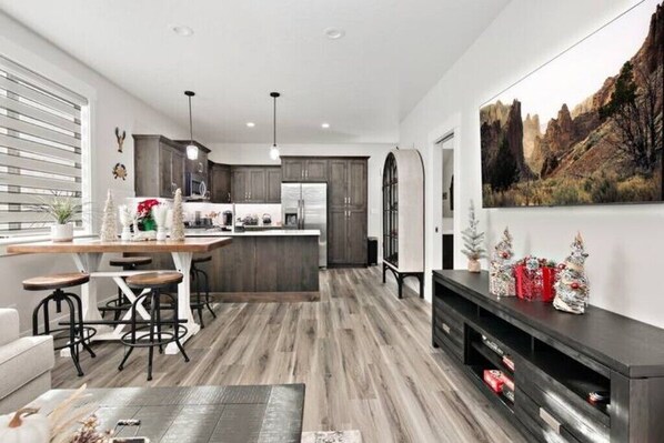 A modern kitchen with dark cabinets, a central island, and bar stools. There is a dining table with chairs, a large painting on the wall, and a fireplace with decorative items.