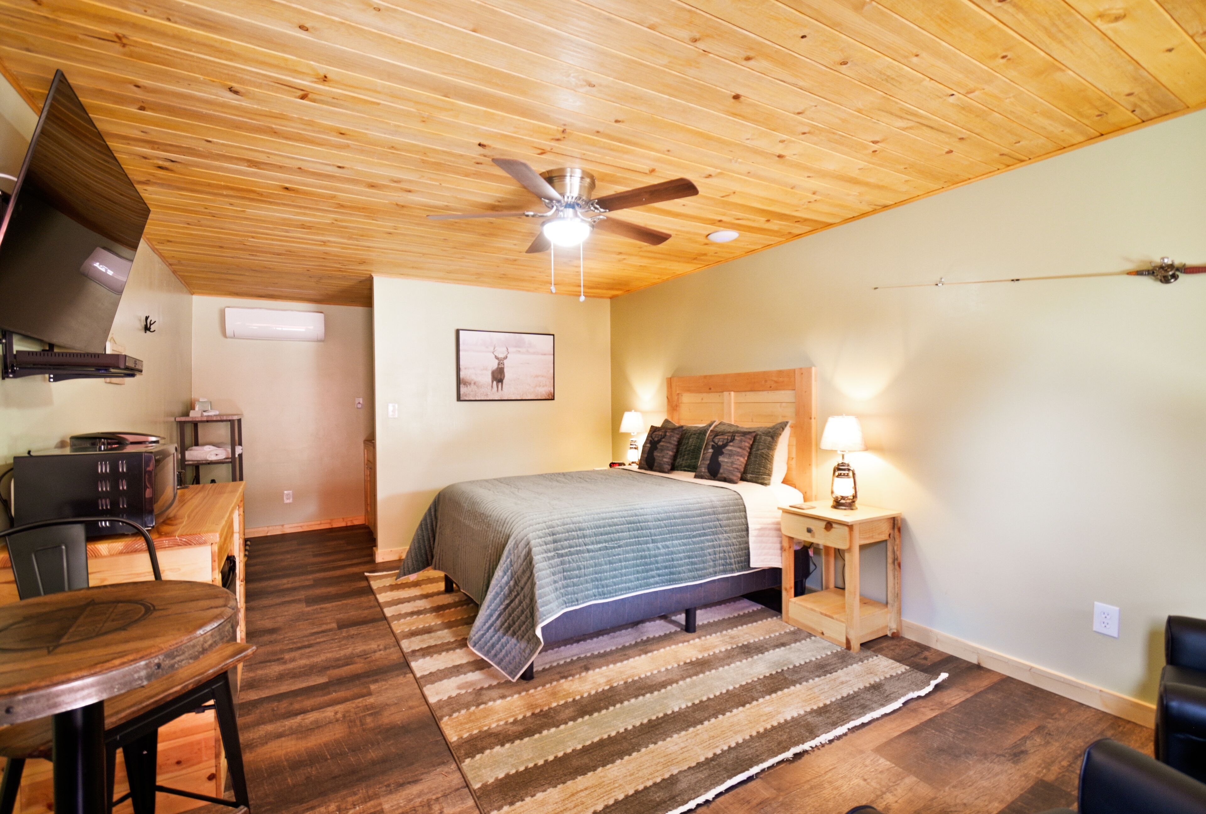 A bedroom with a bed, a desk, a chair, a television, a ceiling fan, and a wall-mounted heater.
