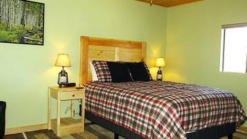A bedroom with a wooden ceiling, a bed with a plaid bedspread, two bedside lamps, a nightstand, a window, and a painting on the wall.