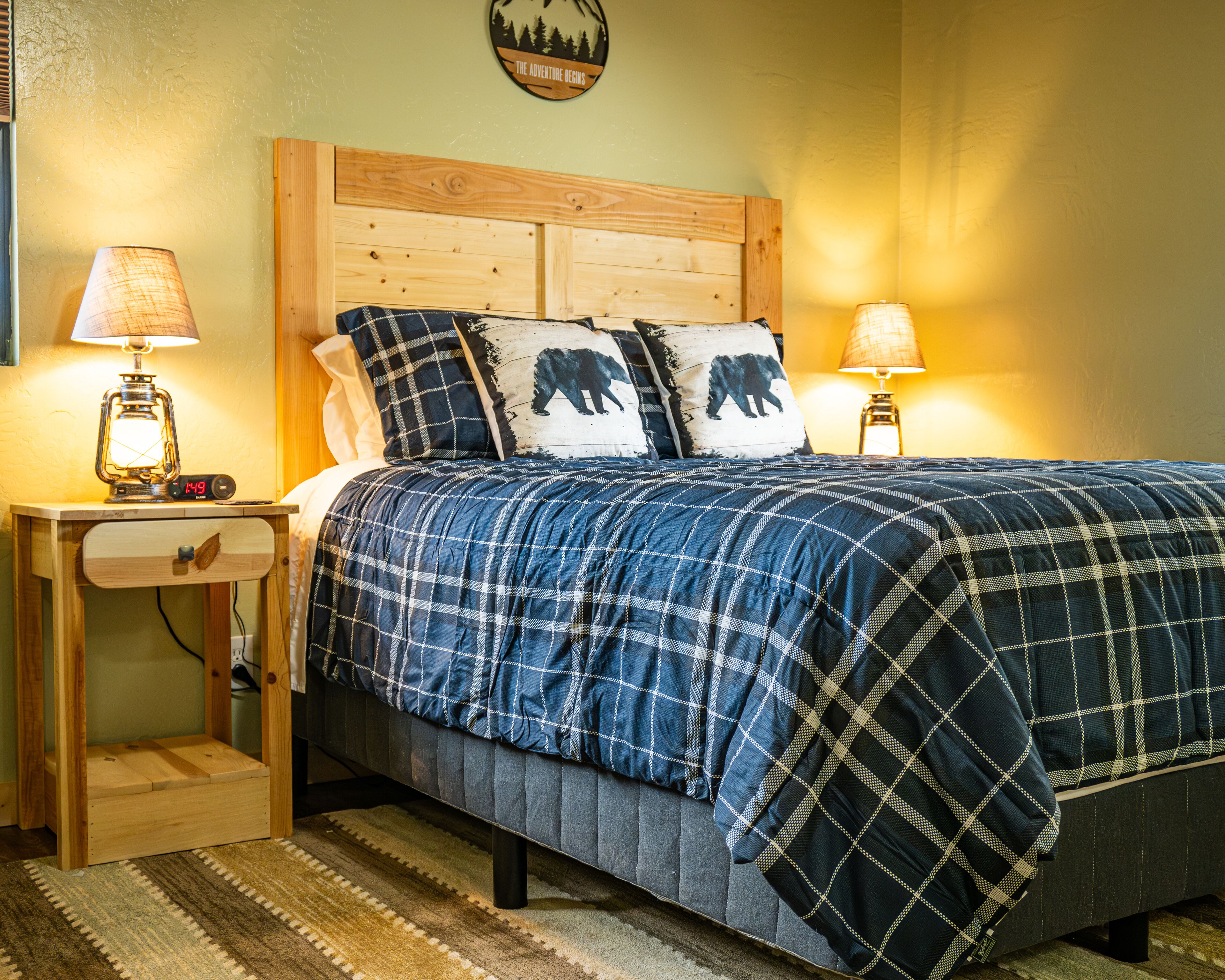 A bedroom with a wooden headboard, a plaid bedspread, two bedside lamps, and a wall clock.