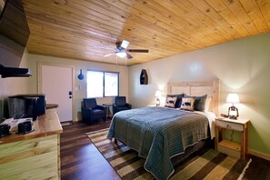 Un dormitorio con techo de madera, una cama con colcha azul, una mesita de noche con lámpara, un escritorio con cafetera y una silla.