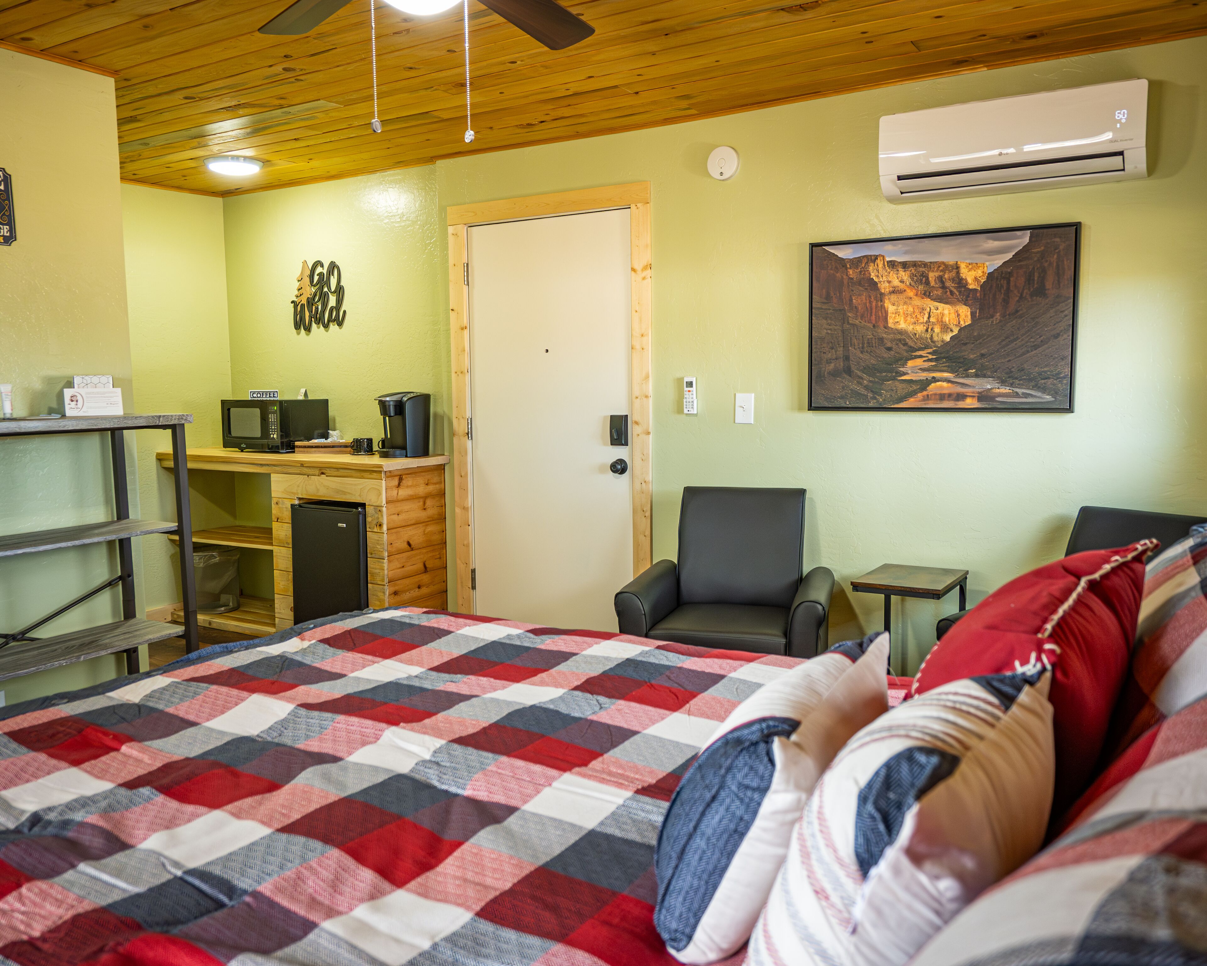 A room with a bed, a desk with a computer, an air conditioner, a chair, and a painting of a canyon.
