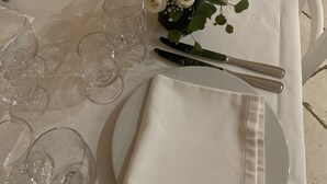 Una llarga taula de menjador coberta amb estovalls blancs, plats i coberts disposats amb cura, i gots de cristall transparent.