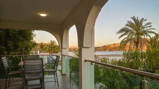 A balcony with two chairs and a table, overlooking a beach and palm trees.