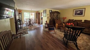 A spacious living room with hardwood floors, a dining area with a wooden table and chairs, a brown leather sofa, and a large wooden cabinet.
