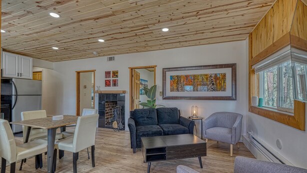 Ein modernes Wohnzimmer mit Holzboden, einem Kamin, einem Essbereich mit Tisch und Stühlen und einem großen Fenster mit Blick auf Bäume.