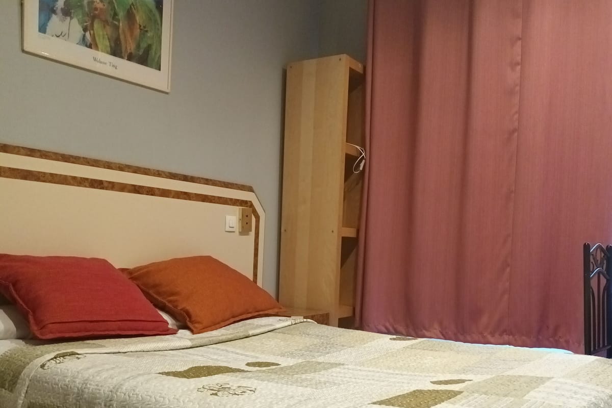 Quarto com cama, travesseiros, colcha, guarda-roupa de madeira, cortina rosa e um quadro na parede.