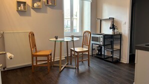 A small, well-lit room with a wooden floor, a round table with two chairs, a radiator, a window with blinds, and a kitchenette area with a microwave and shelves.