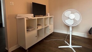 A room with a white entertainment unit, a flat-screen TV, a white standing fan, and a small shelf with towels.