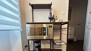 A compact kitchen with a stainless steel refrigerator, a coffee maker, and a small shelf with kitchen essentials.