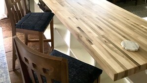 Une longue table à manger en bois, avec des chaises, une plante en pot et une fenêtre laissant entrer la lumière naturelle.