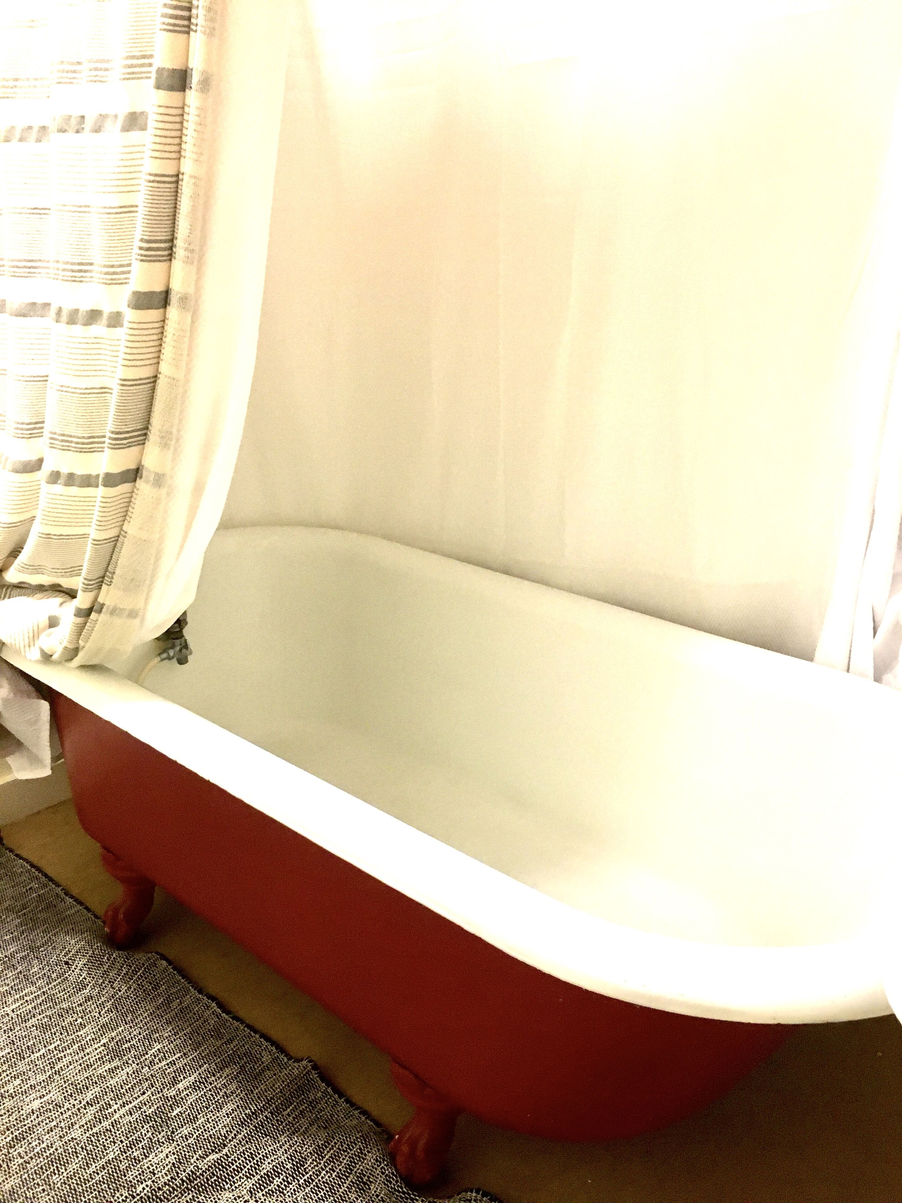 A red clawfoot bathtub with a white interior, a striped curtain partially drawn, and a textured rug on the floor.