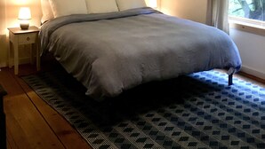 Une chambre à coucher avec un lit, une table de chevet, une lampe, un tableau encadré au mur et une fenêtre donnant sur des arbres.