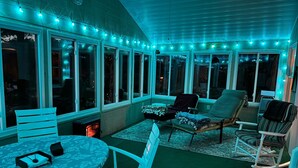 A covered patio with a round table and chairs, a rocking chair, and string lights. - Cassetta's View - Hickory Lake N.Y. (De Peyster)