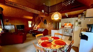 A cozy dining area with a round table set for four, surrounded by wooden chairs. A staircase leads to an upper level, and a kitchen is visible in the background. - Cassetta's View - Hickory Lake N.Y. (De Peyster)