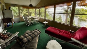A room with a green carpet, a red couch, a table with chairs, a ceiling fan, and large windows overlooking a body of water. - Cassetta's View - Hickory Lake N.Y. (De Peyster)