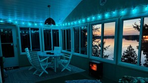 A sunroom with a view of a lake, a table with chairs, and a fireplace. - Cassetta's View - Hickory Lake N.Y. (De Peyster)