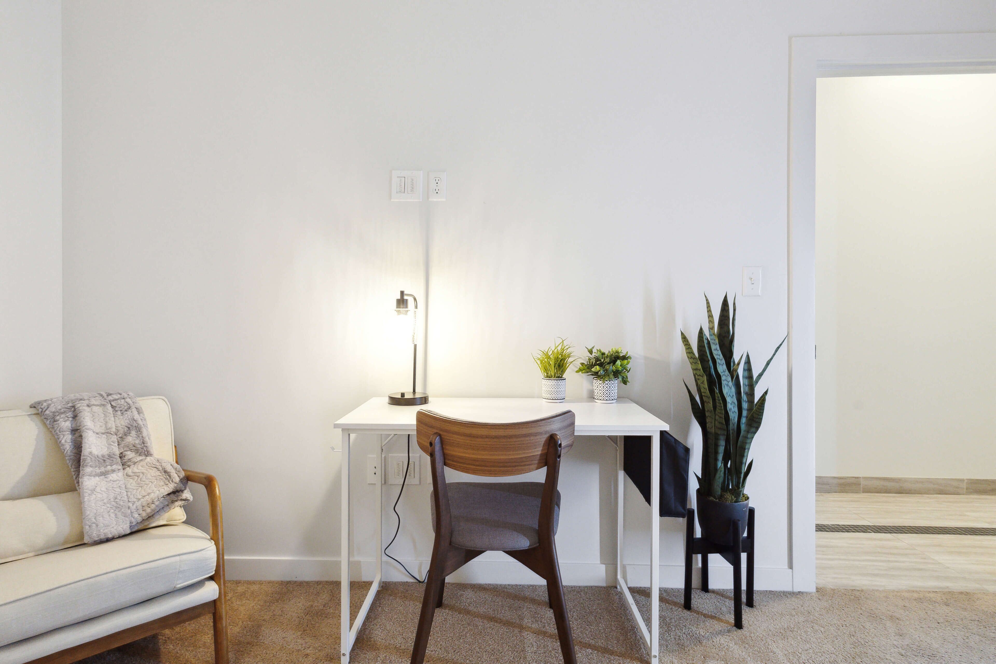 Un espacio de oficina minimalista con un escritorio blanco, una silla de madera, una lámpara y dos plantas en macetas.