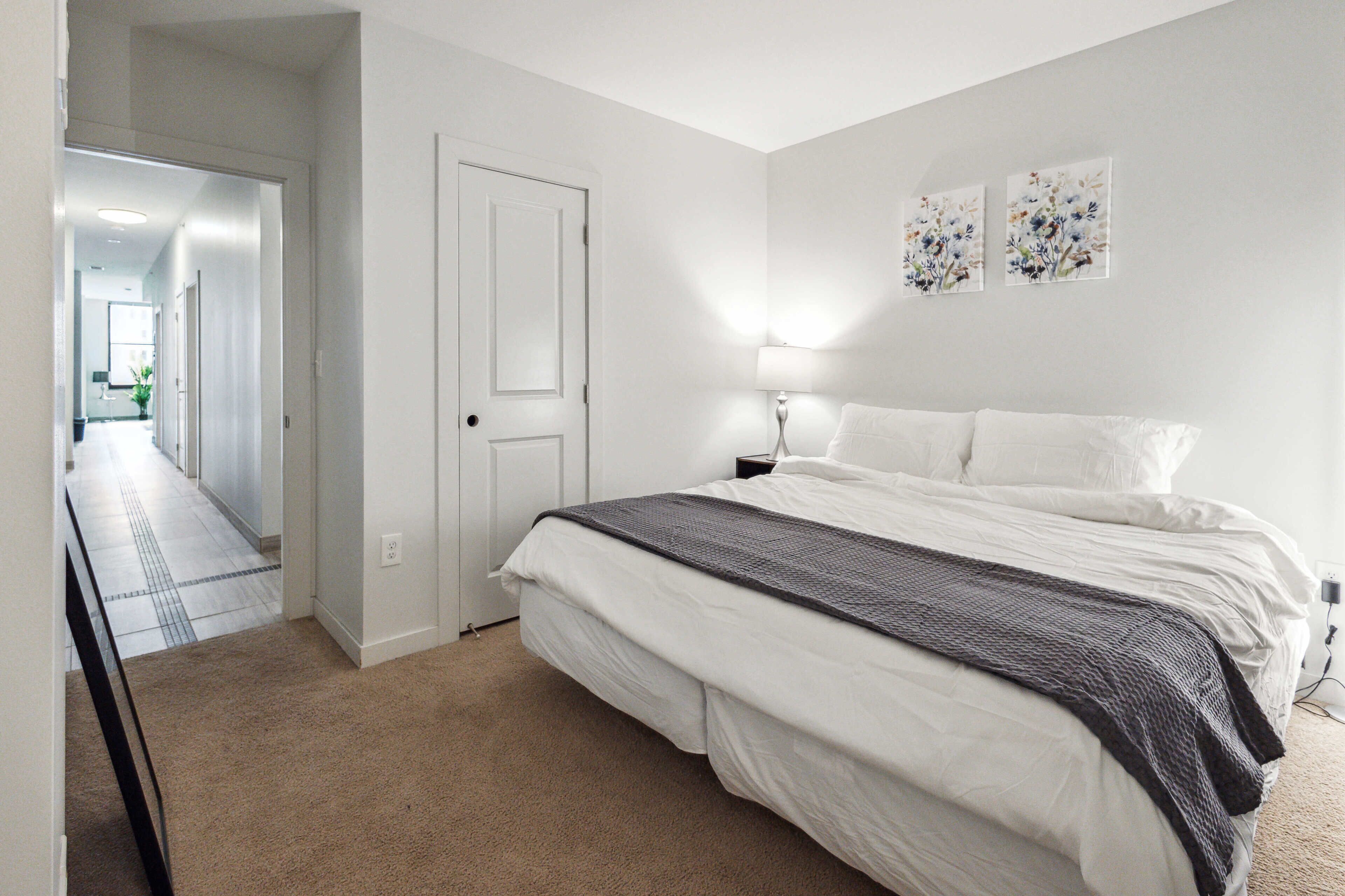 A bedroom with a large bed, a bedside table, a lamp, and a door leading to a hallway.
