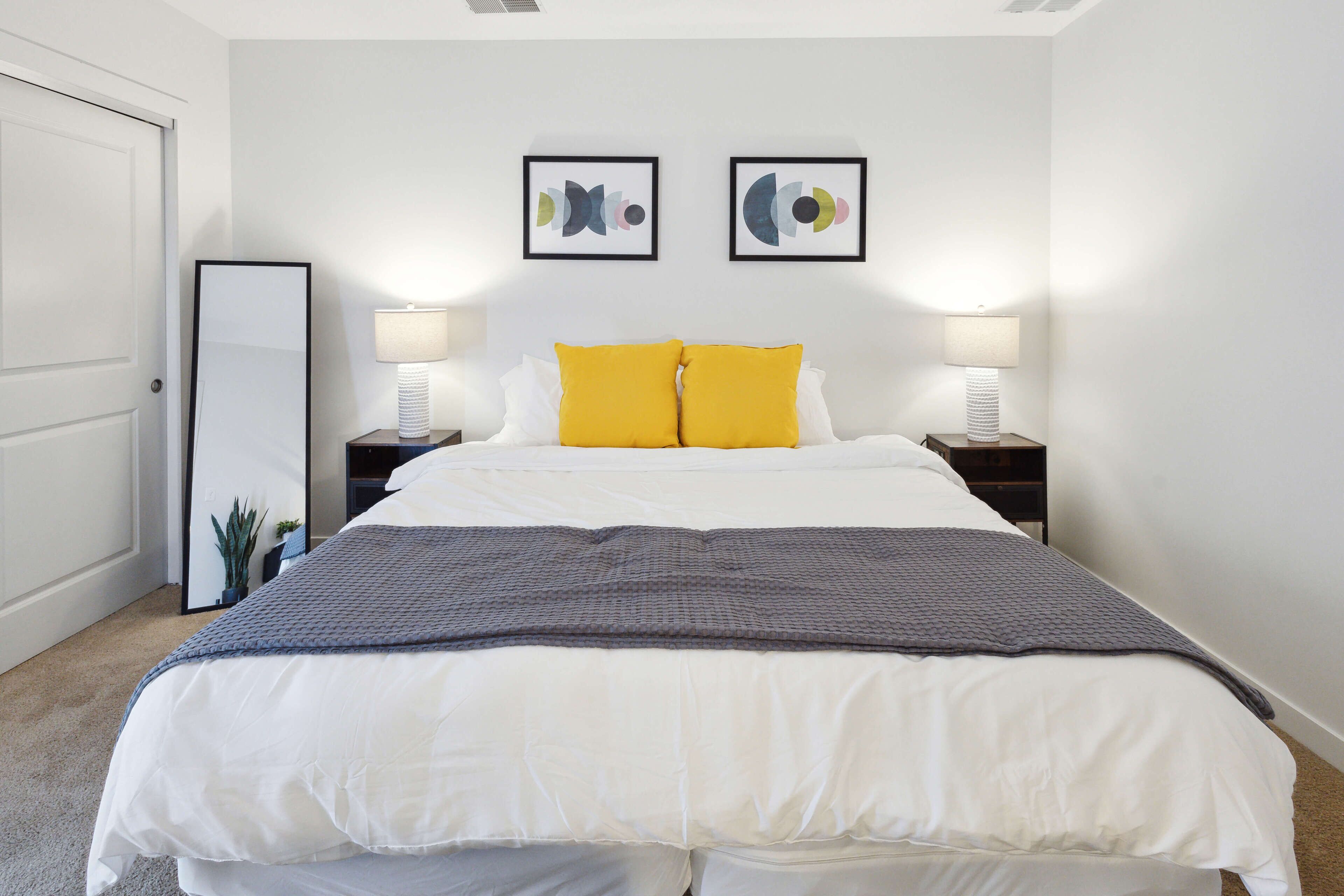 A neatly made bed with white and gray bedding, two bedside lamps, and two abstract art pieces on the wall.
