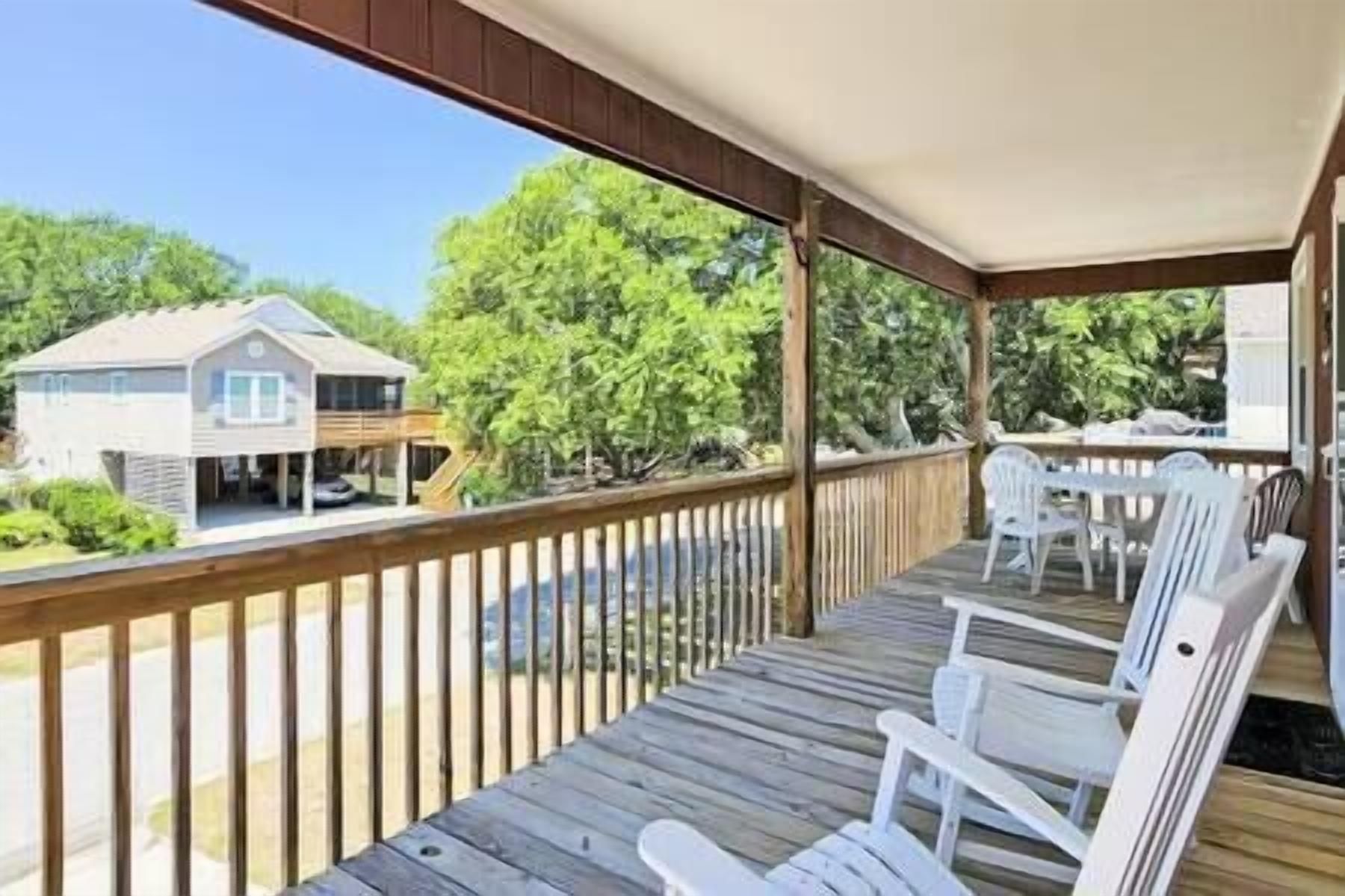 A covered wooden deck with white plastic chairs, a view of a house, and a wooden railing.