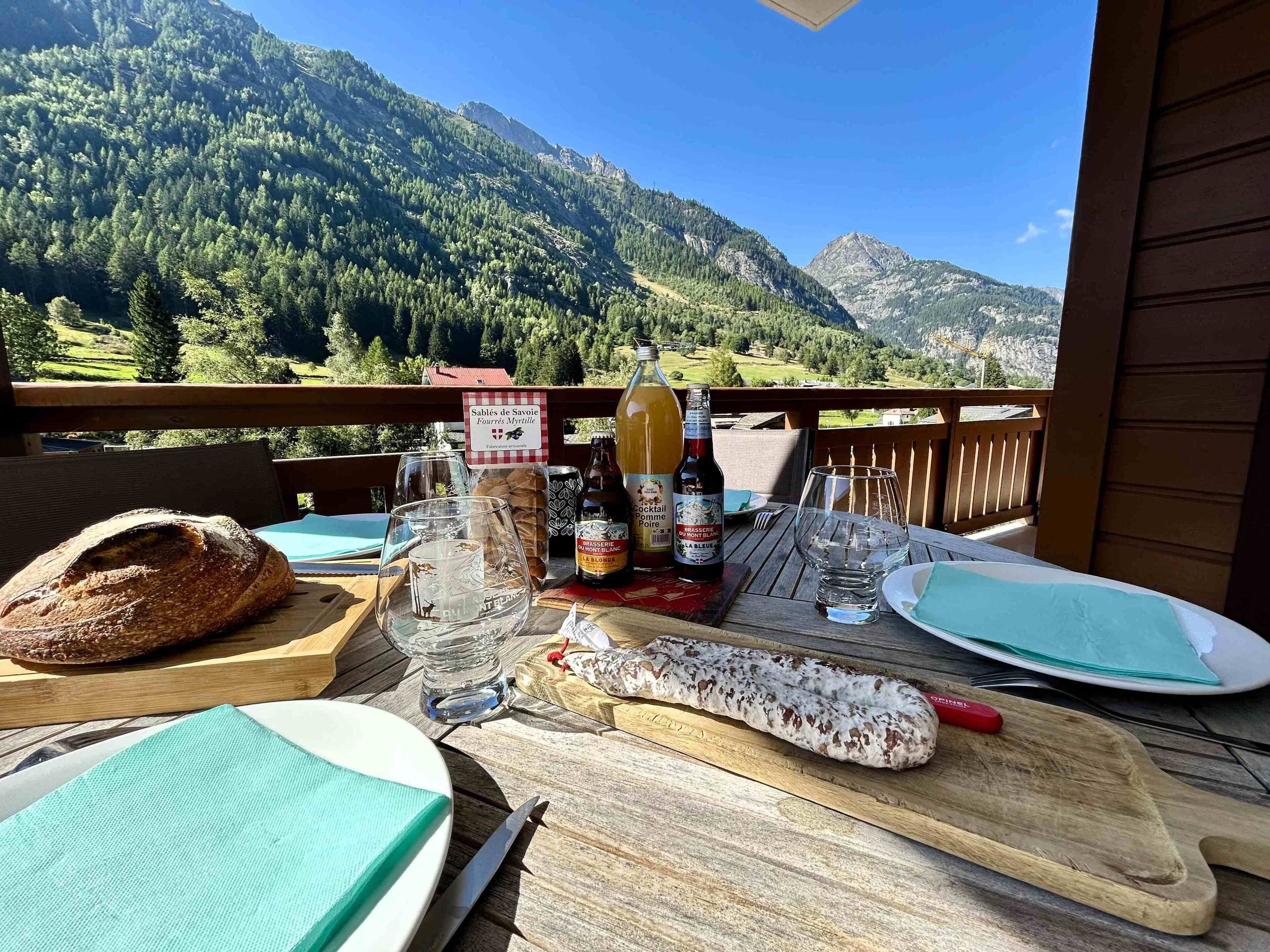 A table set outdoors with a mountain view, featuring a loaf of bread, a bottle of beer, a glass pitcher, and a bottle of wine.