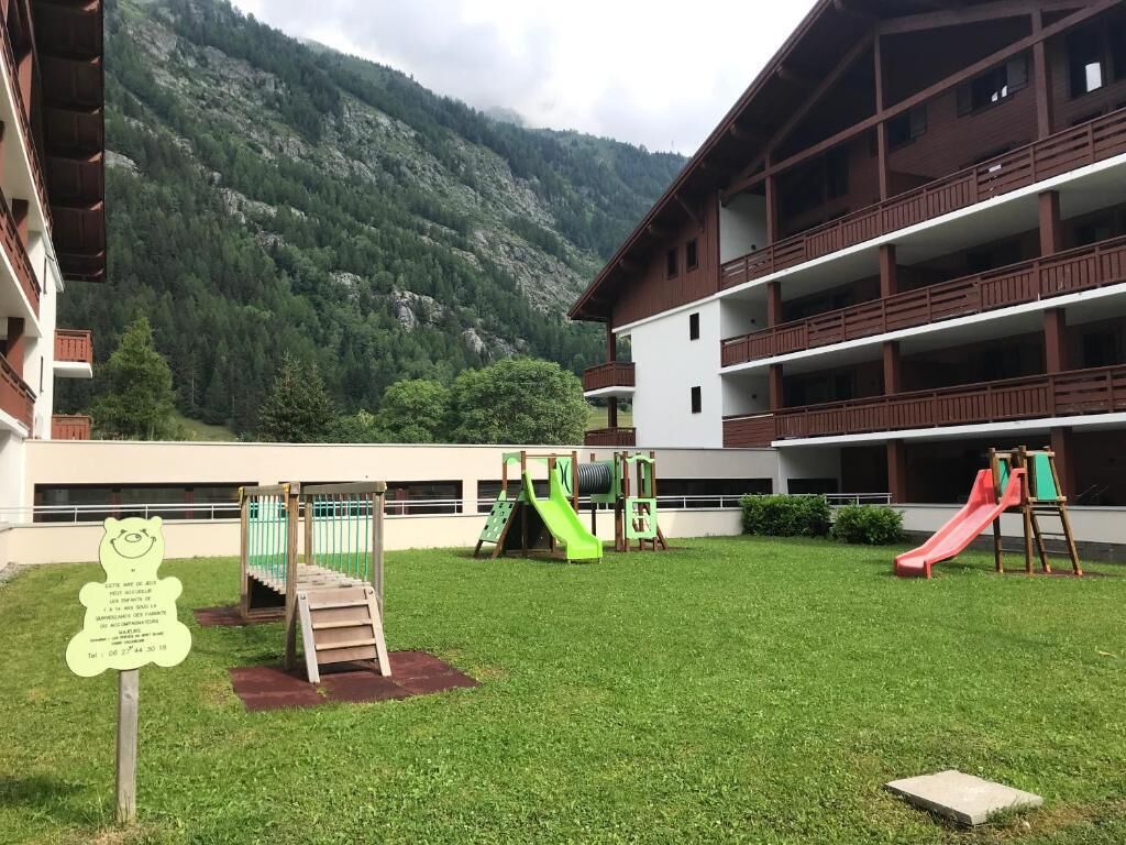 Un terrain de jeux avec des toboggans et des structures d’escalade, entouré d’un bâtiment doté de balcons, le tout sur fond de paysage montagneux.