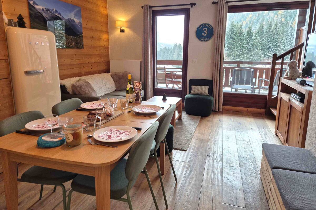 A cozy room with a wooden dining table set for a meal, a sofa, and a view of a snowy forest through a large window.