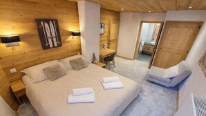 A hotel room with a wooden ceiling, a large bed with two pillows, a bedside table, a chair, a desk, and a mirror.