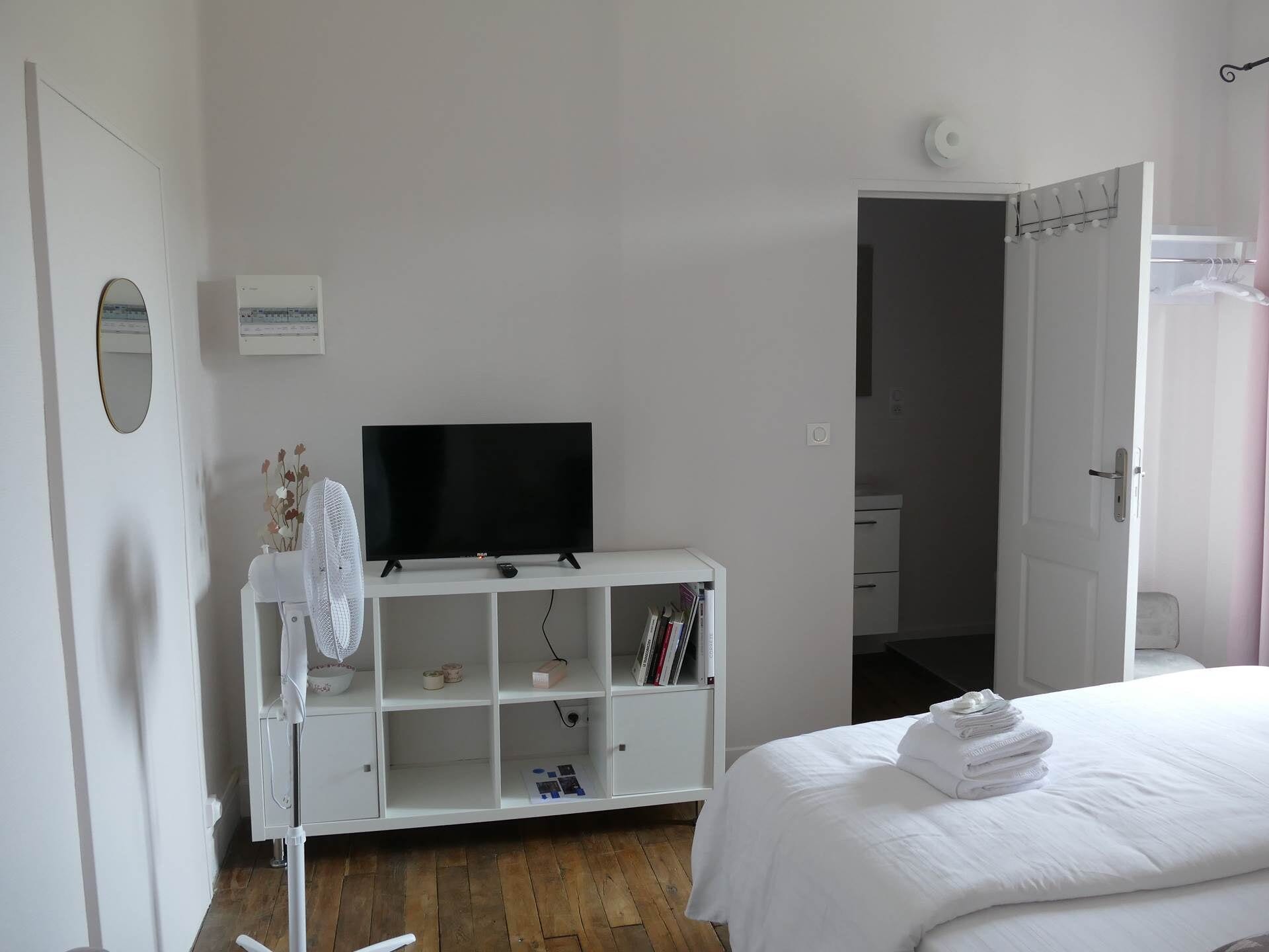 A bedroom with a white bed, a TV on a white stand, a white wardrobe, and a door leading to another room.