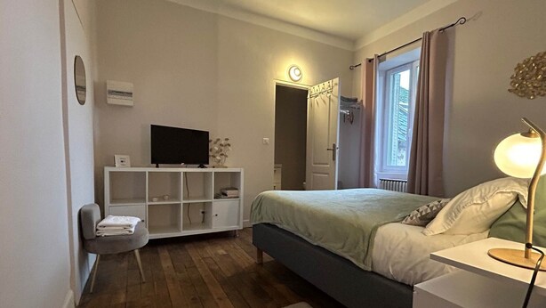 Ein Schlafzimmer mit Bett, Nachttisch, Lampe, Fernseher und einer kleinen Sitzecke mit Sessel und Kissen.
