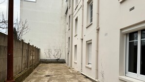 A narrow alleyway with a concrete floor, a red pole, and a white building with multiple windows and vents. - L'escale (Rennes)