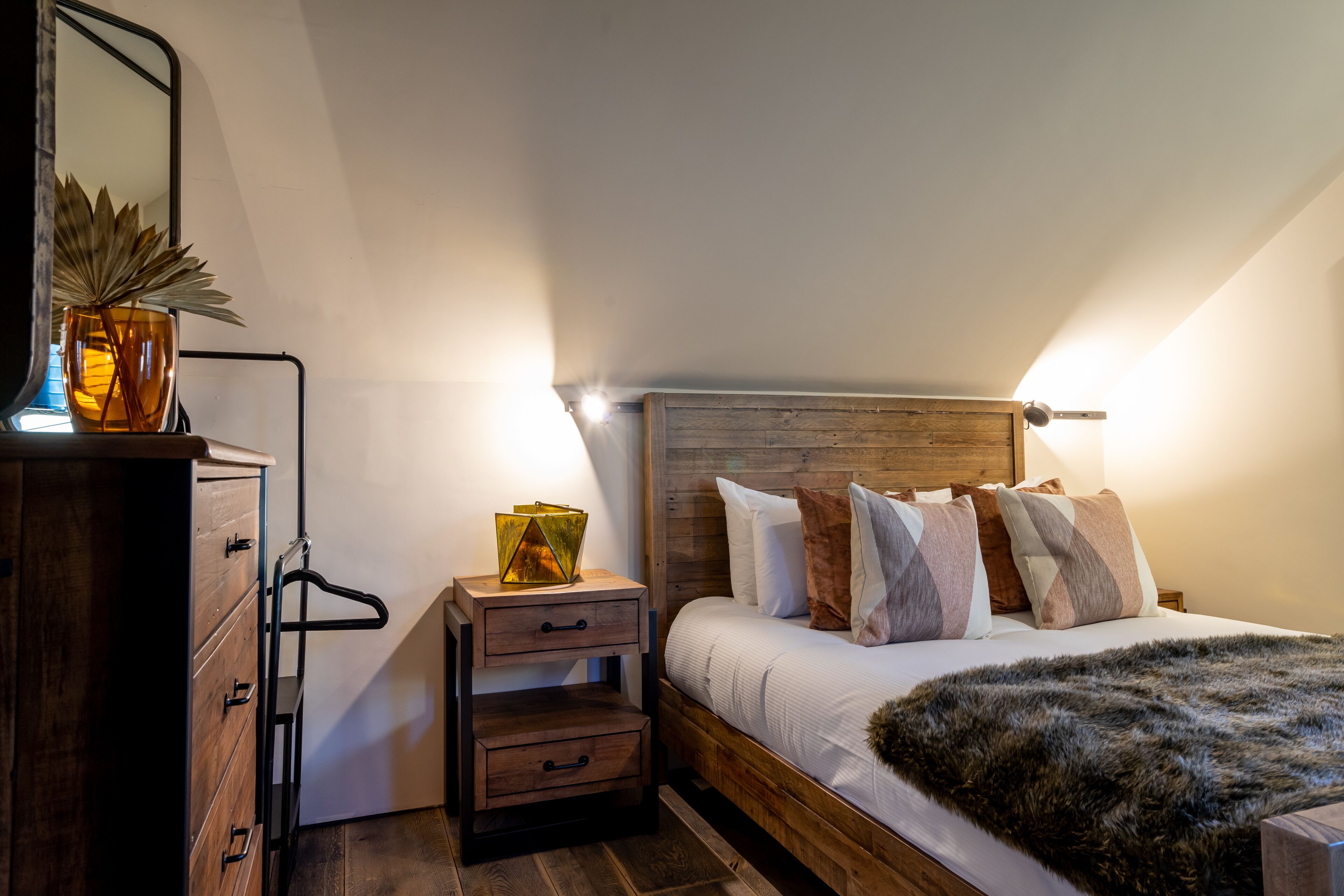 A bedroom with a wooden headboard, a bed with multiple pillows, a nightstand with a lamp, and a dresser with drawers.