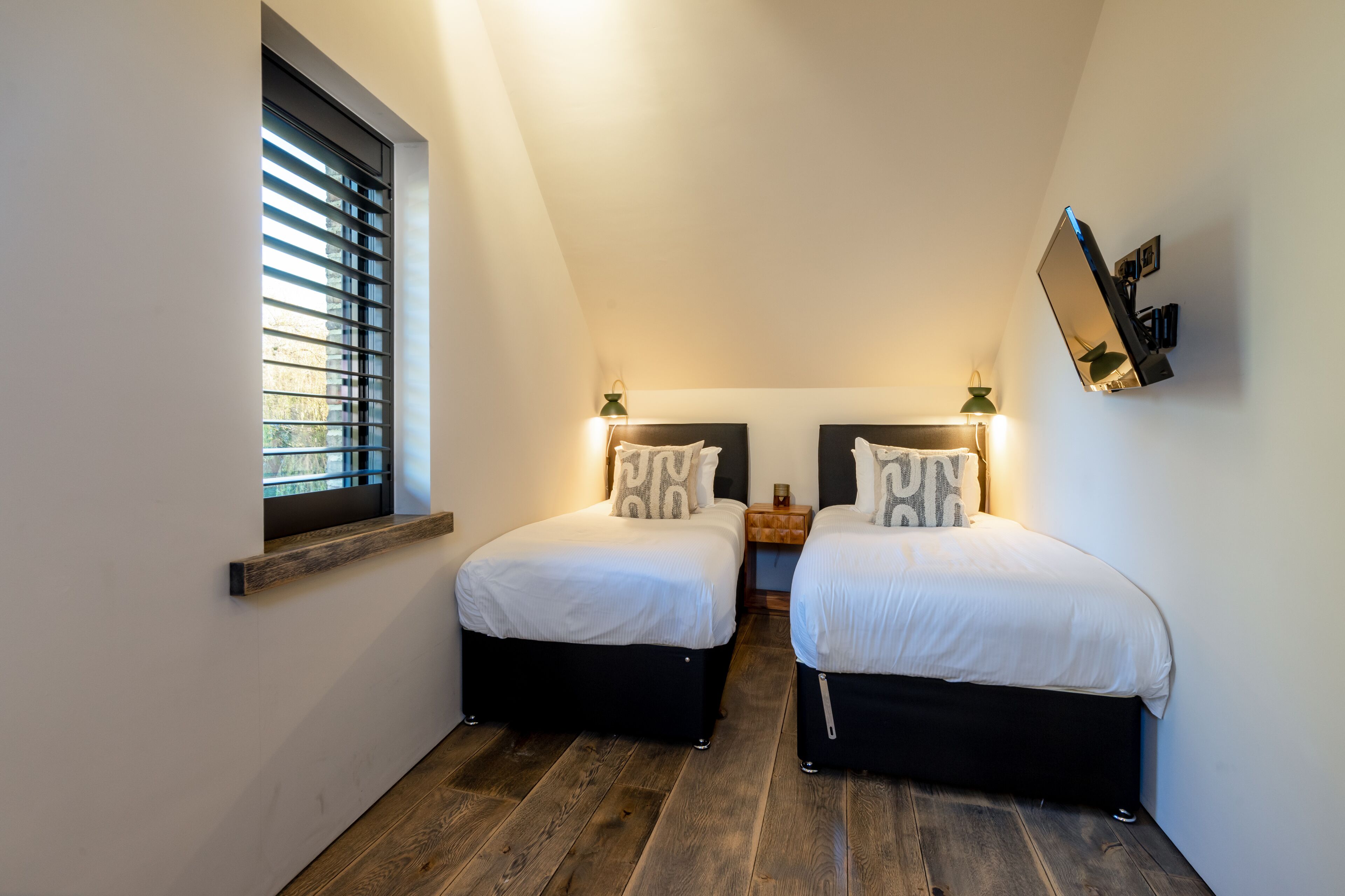 Een slaapkamer met twee bedden, een raam met jaloezieën, bedlampen en een aan de muur bevestigde televisie.