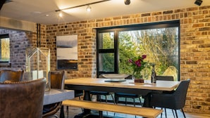 Un coin repas au style rustique, avec un mur en briques, une table en bois, des chaises en cuir et une grande fenêtre donnant sur un paysage verdoyant.