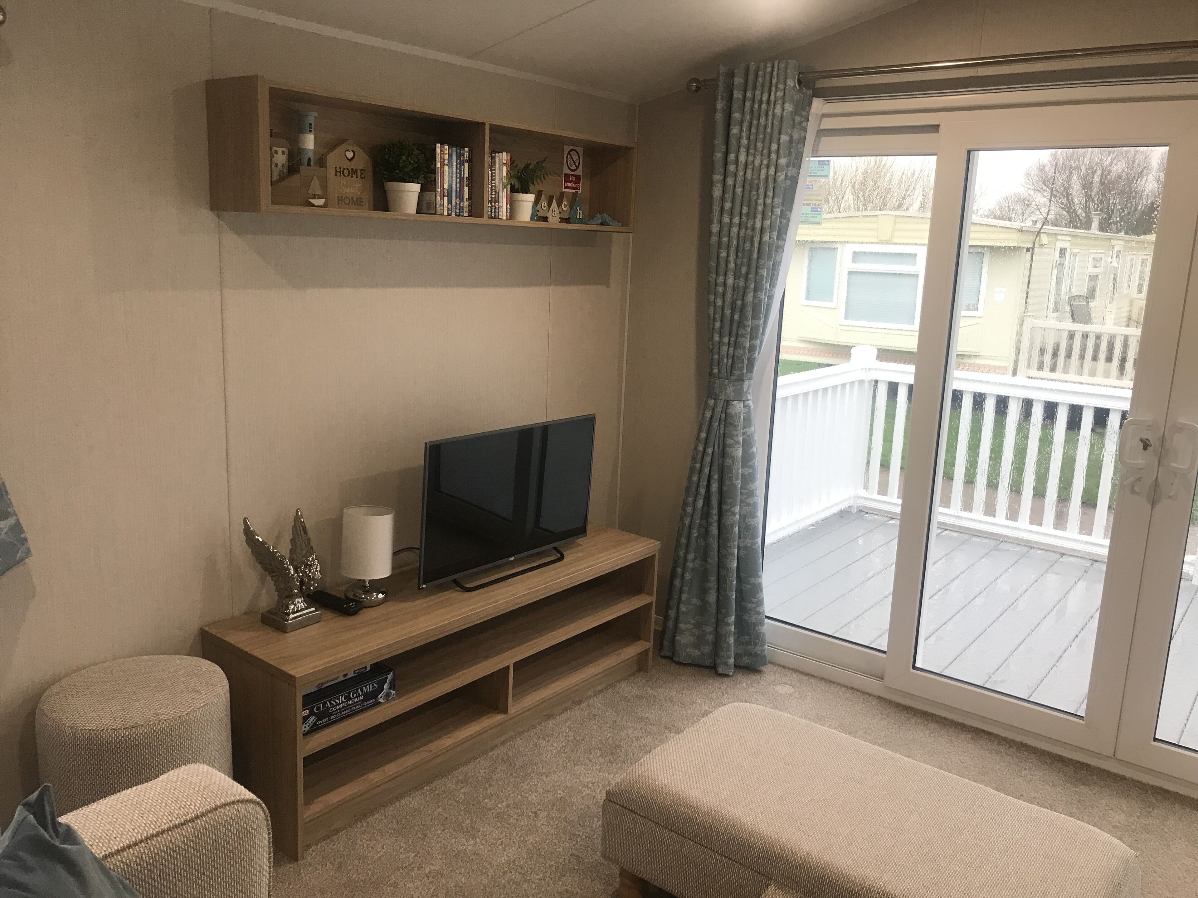A living room with a flat-screen TV on a wooden stand, a small table with a lamp, a bookshelf with various items, and a sliding glass door leading to a balcony.