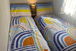A room with two single beds, each with colorful striped bedding. There is a lamp on a bedside table between the beds.