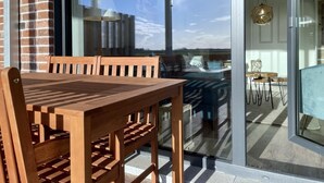 Una mesa de comedor de madera con sillas, un vidrio corredizo que da a un balcón con vista al cielo y una lámpara colgante.