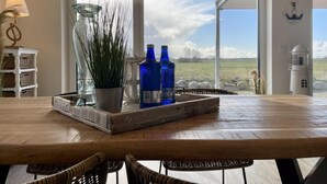 Un comedor con una mesa de madera, sillas de mimbre, una bandeja con una planta en maceta y botellas azules, y vistas al campo a través de amplias ventanas.