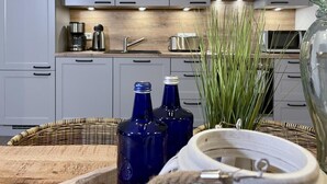 Una cocina con armarios grises, un lavamanos de acero inoxidable y un microondas. Hay dos botellas azules sobre la encimera, una cesta de mimbre y un tazón blanco.