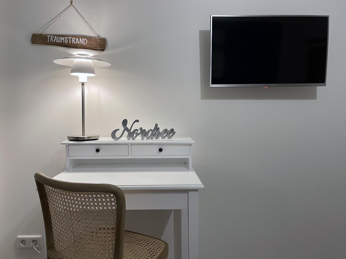A white desk with a lamp, a TV mounted on the wall, and a chair with a woven backrest.