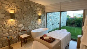 A modern bedroom with a stone wall, a large window, a bathtub, and a bed with a tray of toiletries. - Flat Ilê de Pipa Samoa - By Almare (Tibau do Sul)